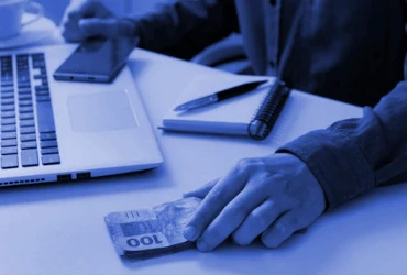 Imagem de uma pessoa usando um computador, um celular, uma agenda e manuseando dinheiro.